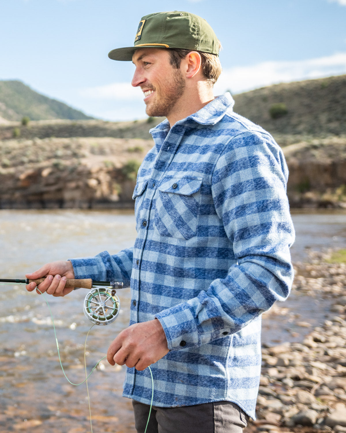 Man in a blue plaid shirt and green cap fly fishing by a river.