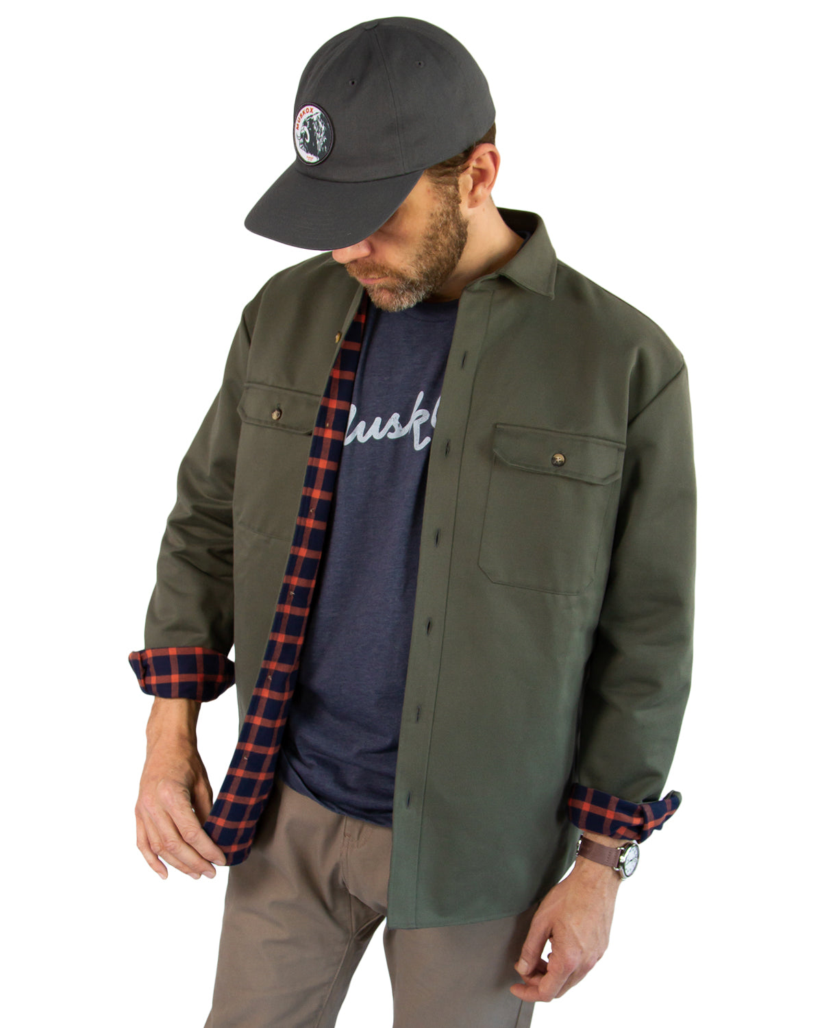 Man wearing a green jacket, navy shirt with plaid sleeves, and gray cap on a white background